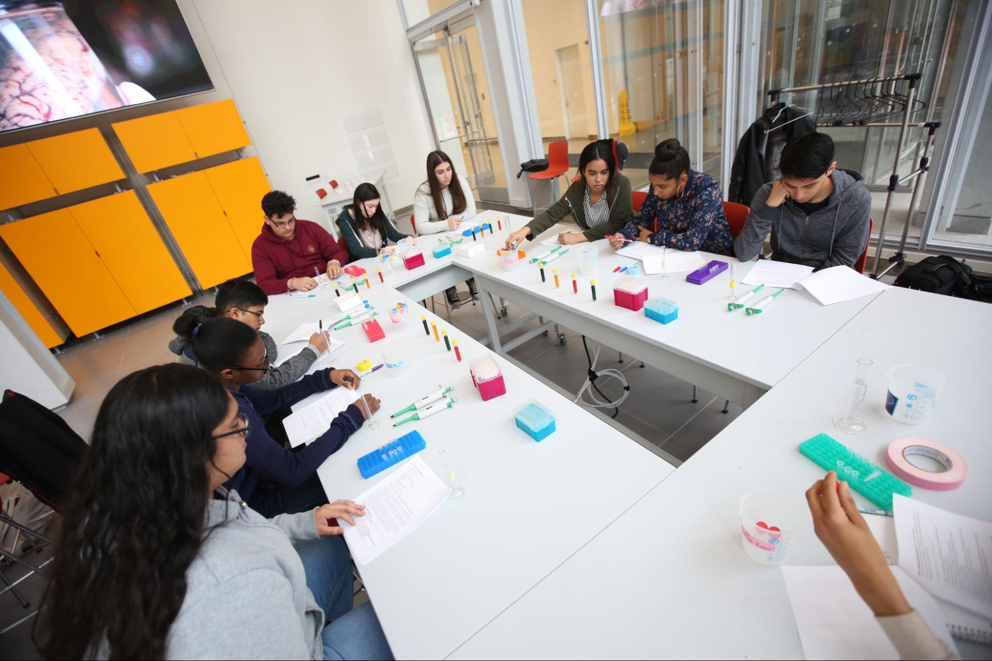 Nine high school students seated around a u-shaped table with small measuring instruments, preparing for a scientific experiment