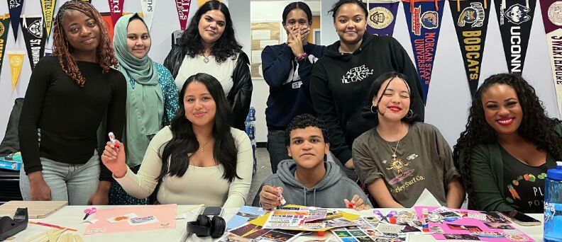Five high school students stand behind three students and an instructor at a table with college banners behind them